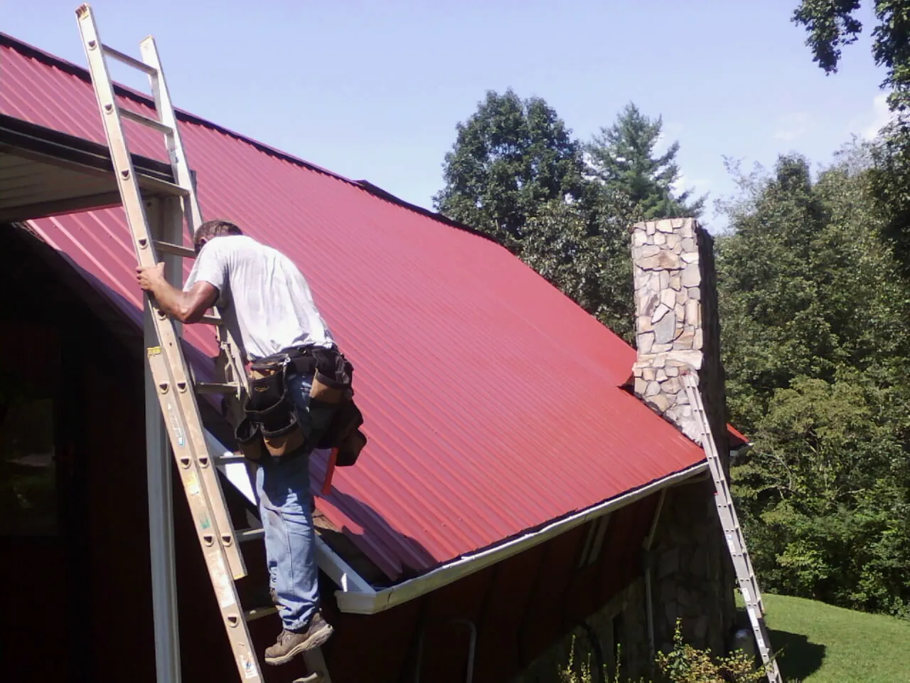 Roofer on a ladder beside a red metal roof and stone chimney — Soffit & Fascia Repair by Roofing Brothers in Mahopac Falls